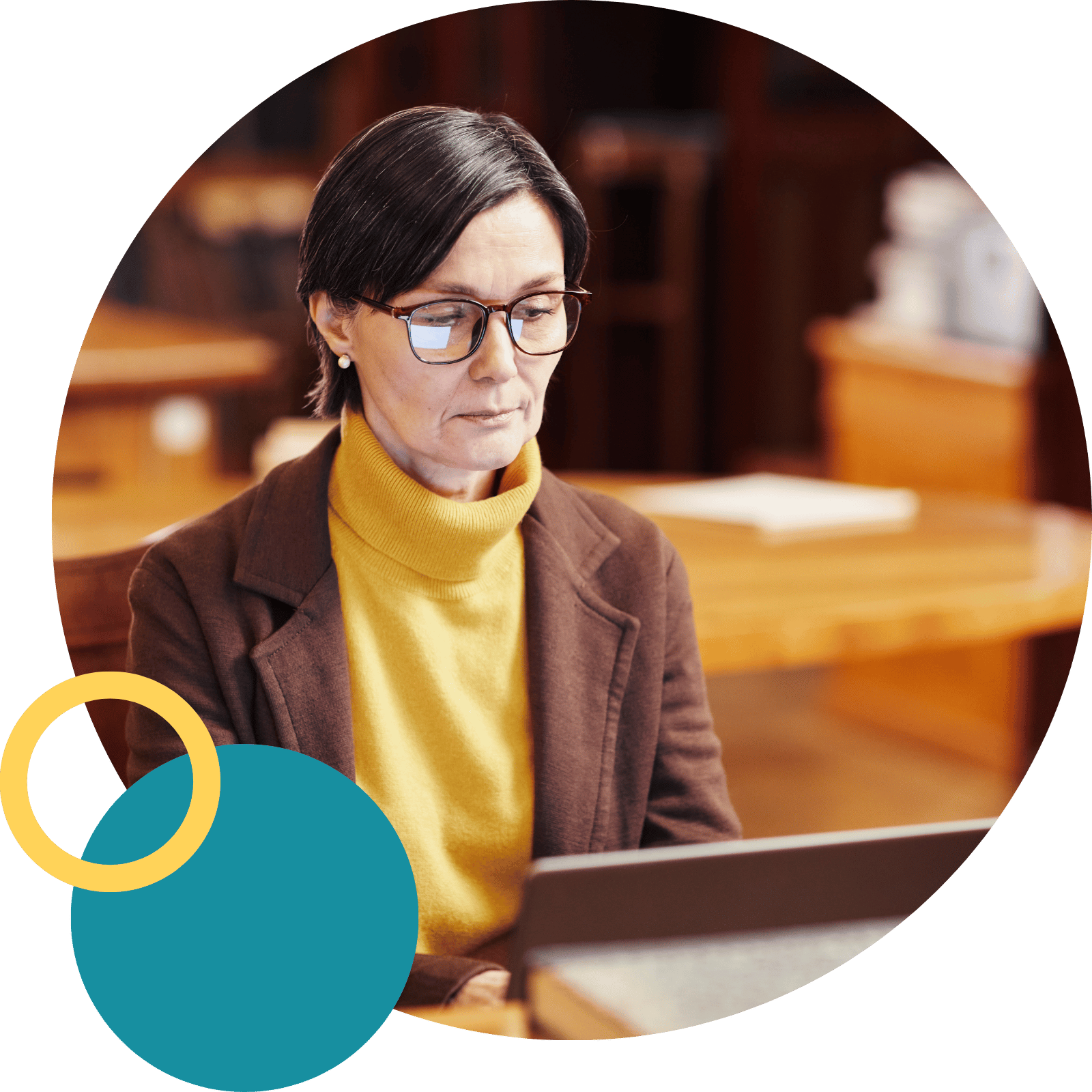 A professional woman with dark hair and glasses is seated at a table, intensely focused on working on her laptop. She is wearing a brown jacket over a mustard yellow turtleneck sweater.